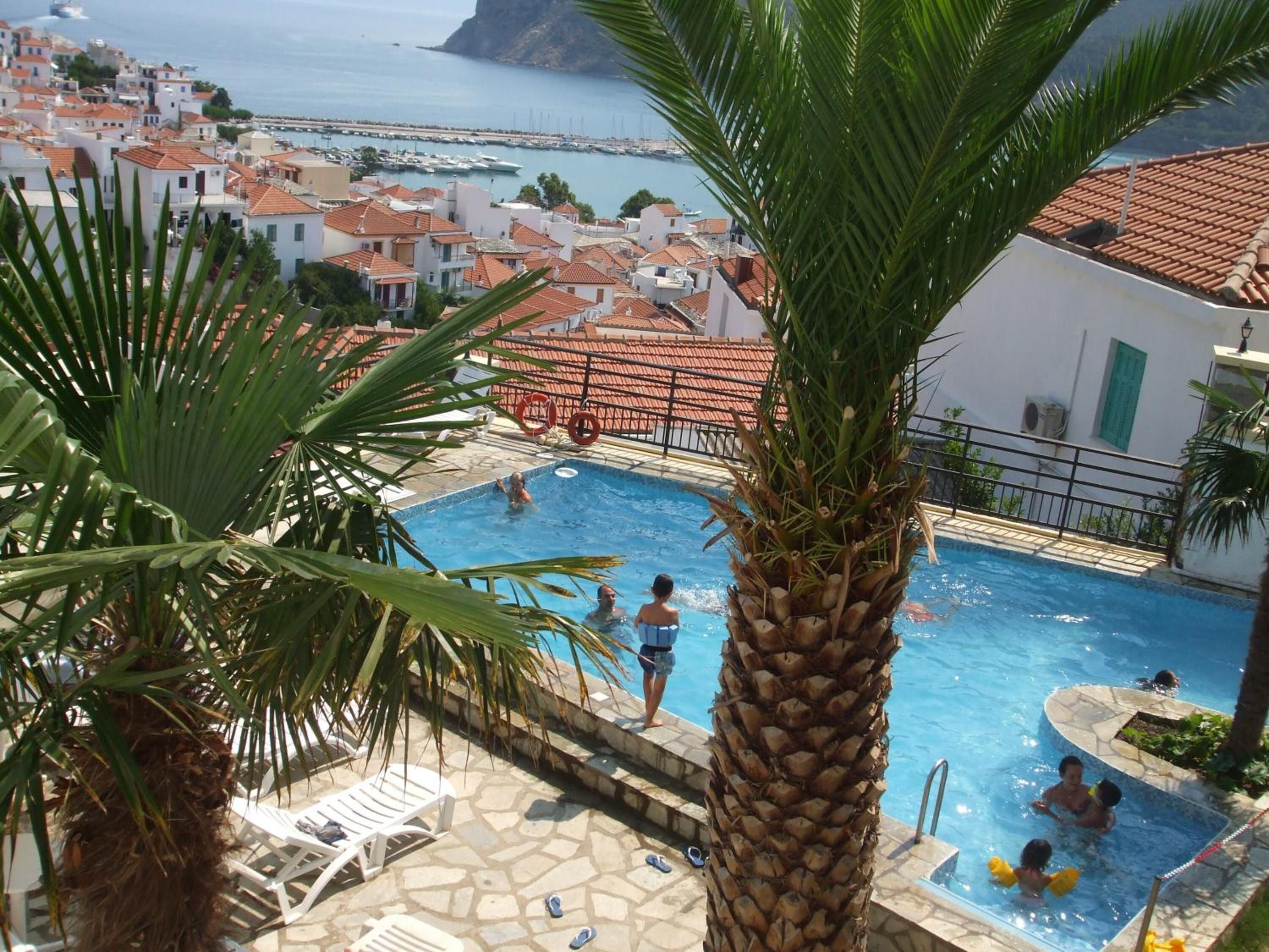 A pool area with palm trees, a view of the sea, and buildings in the background.