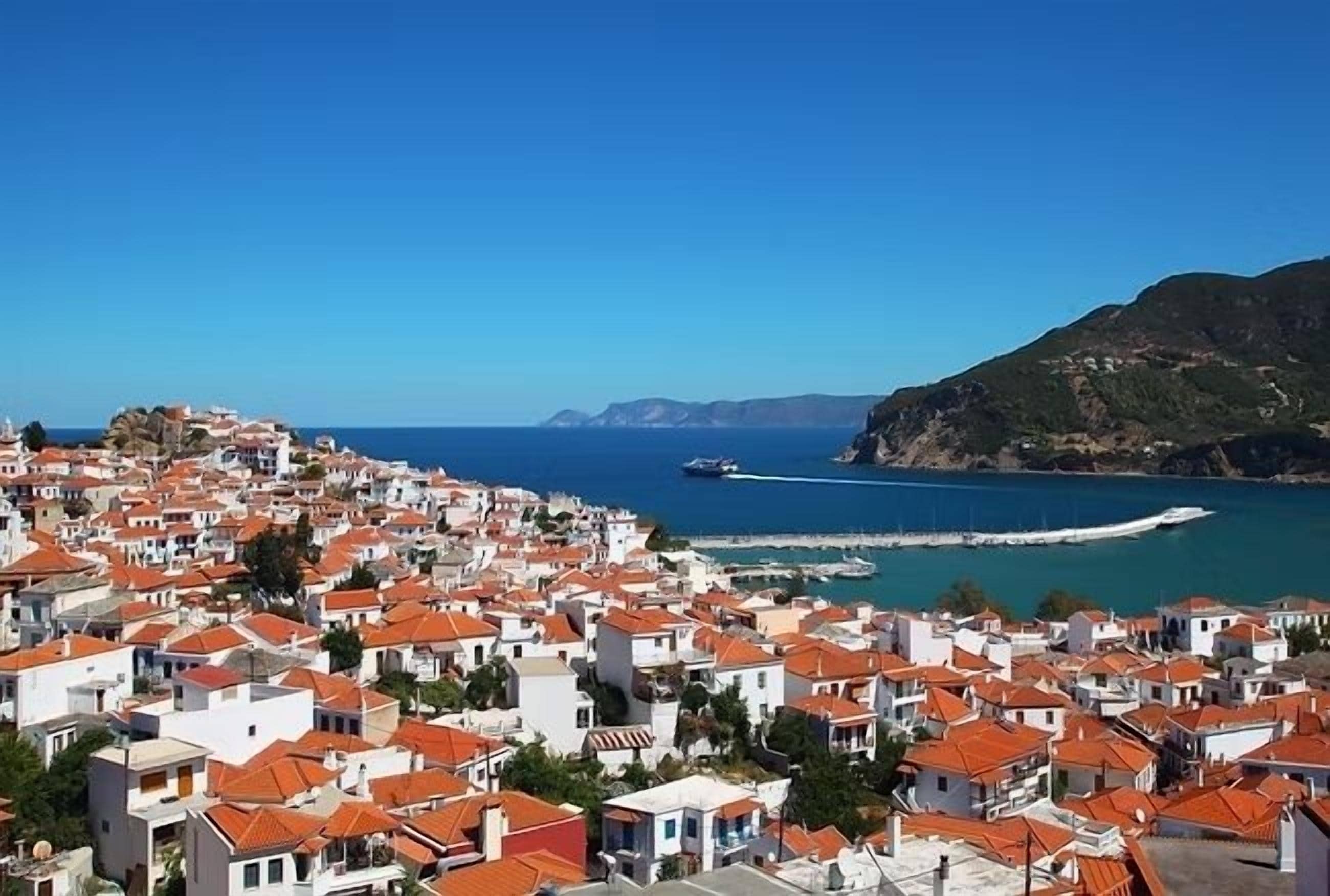 A coastal town with tightly packed buildings, a clear blue sky, and a view of the sea and distant mountains.