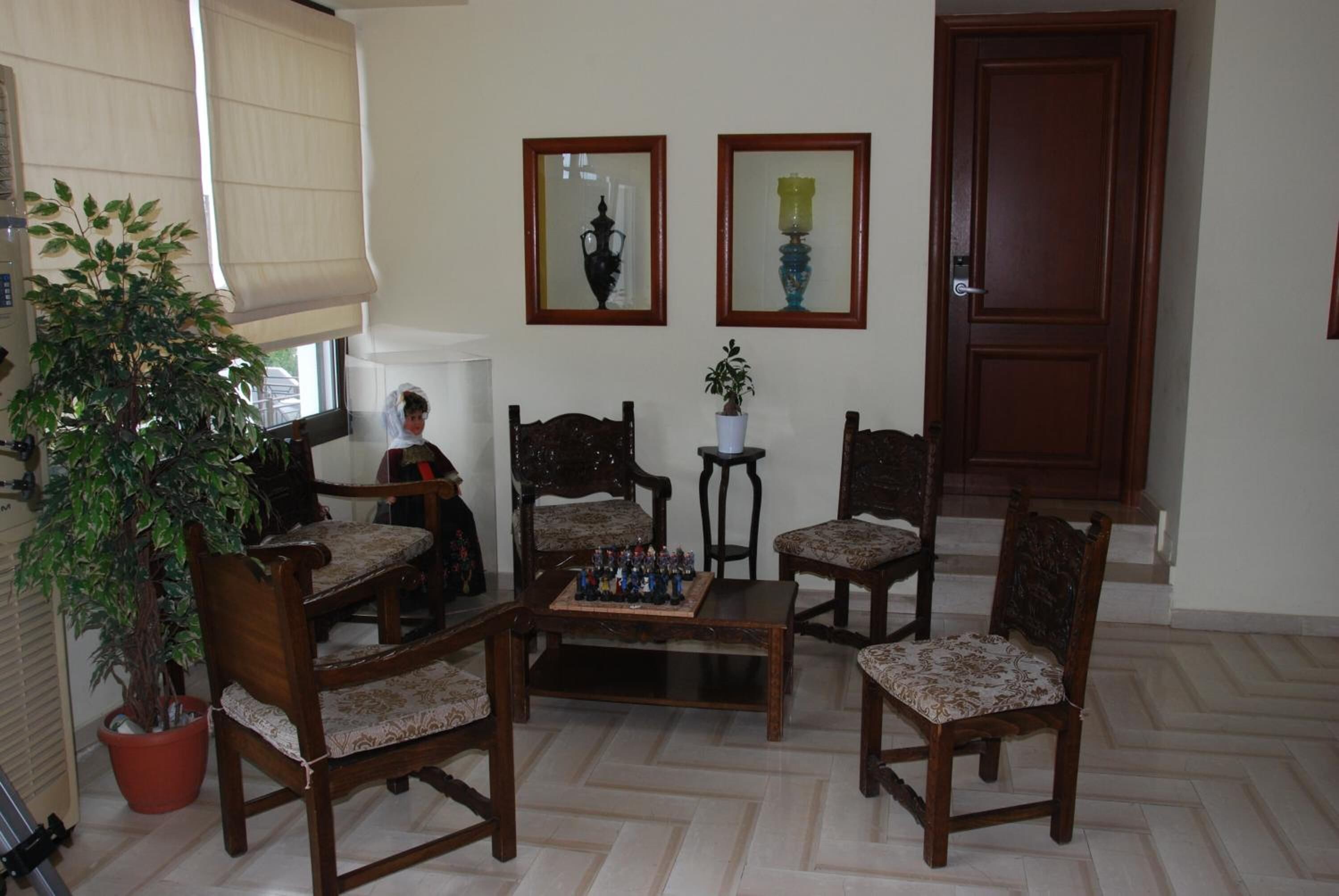A living room with a chess table, four wooden chairs, two potted plants, a large window with blinds, and a door.