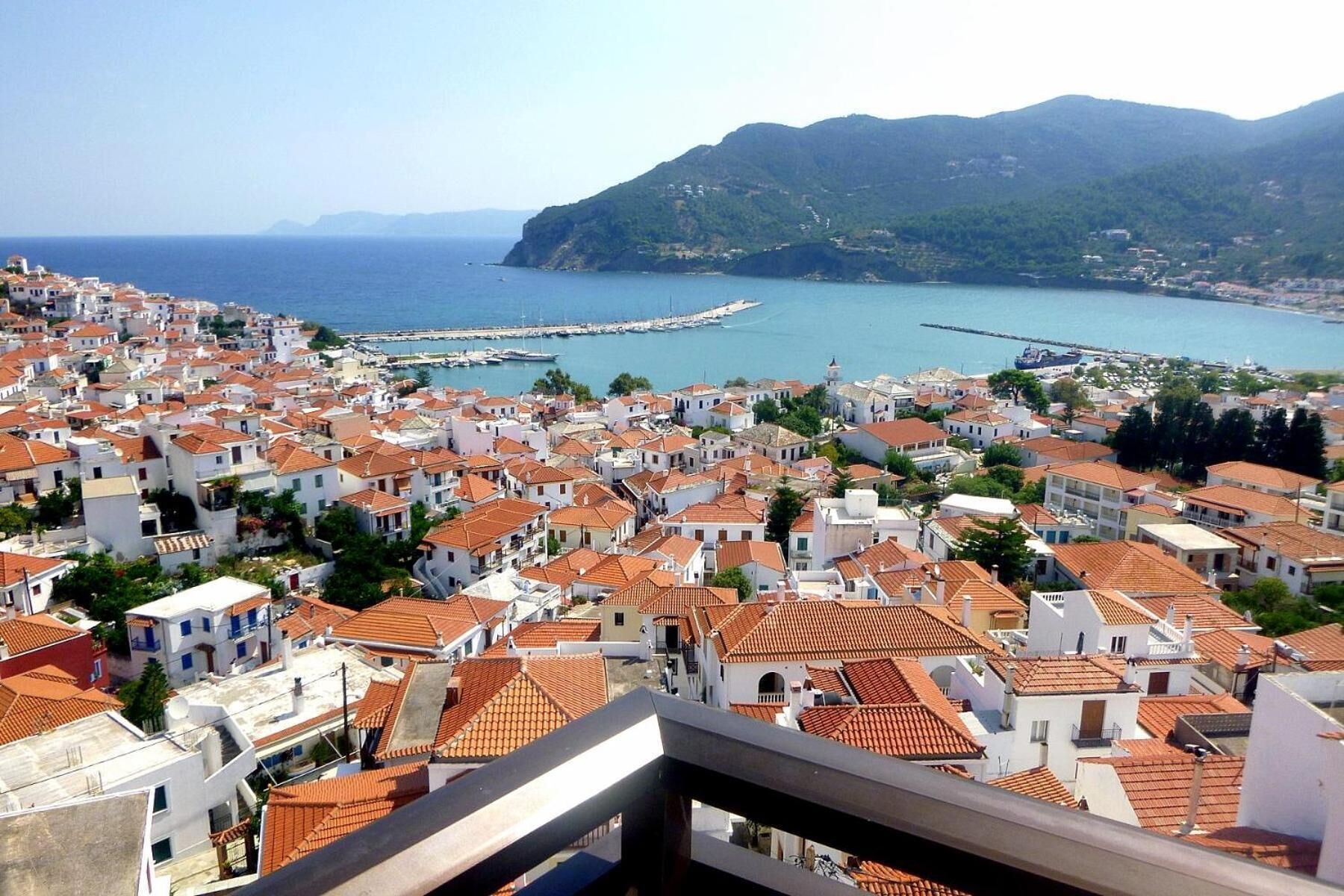 A coastal town with white buildings and red-tiled roofs, a marina, and a mountainous backdrop.
