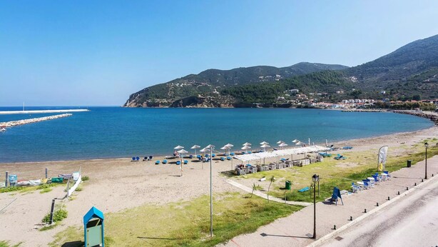 A coastal beach with umbrellas and lounge chairs, a clear sky, and a mountainous backdrop.