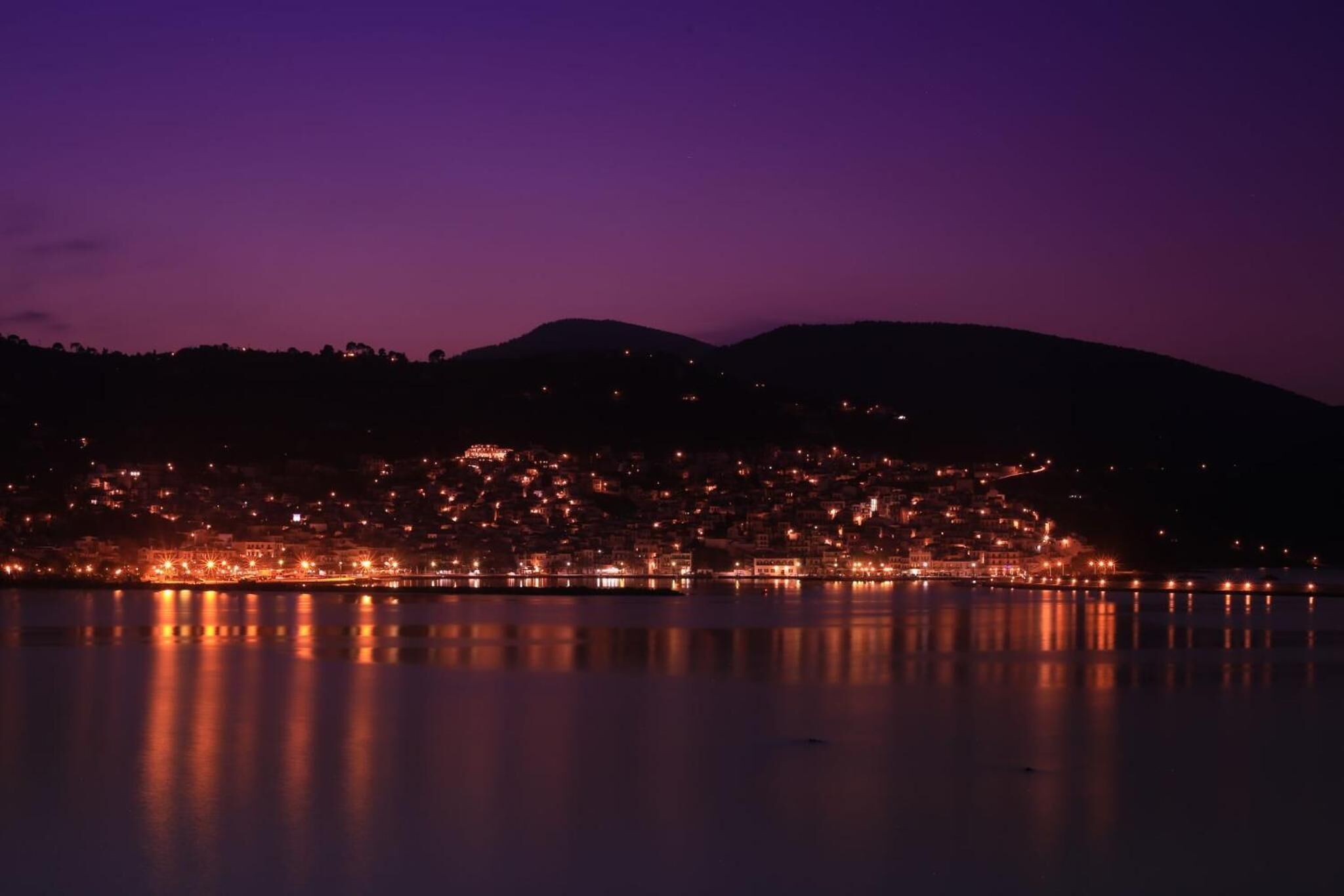 A nighttime cityscape with a calm lake reflecting lights, a hilly backdrop, and a densely populated area with buildings.