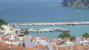 A coastal town with a marina filled with boats, white buildings with red-tiled roofs, and a rocky cliff in the background.