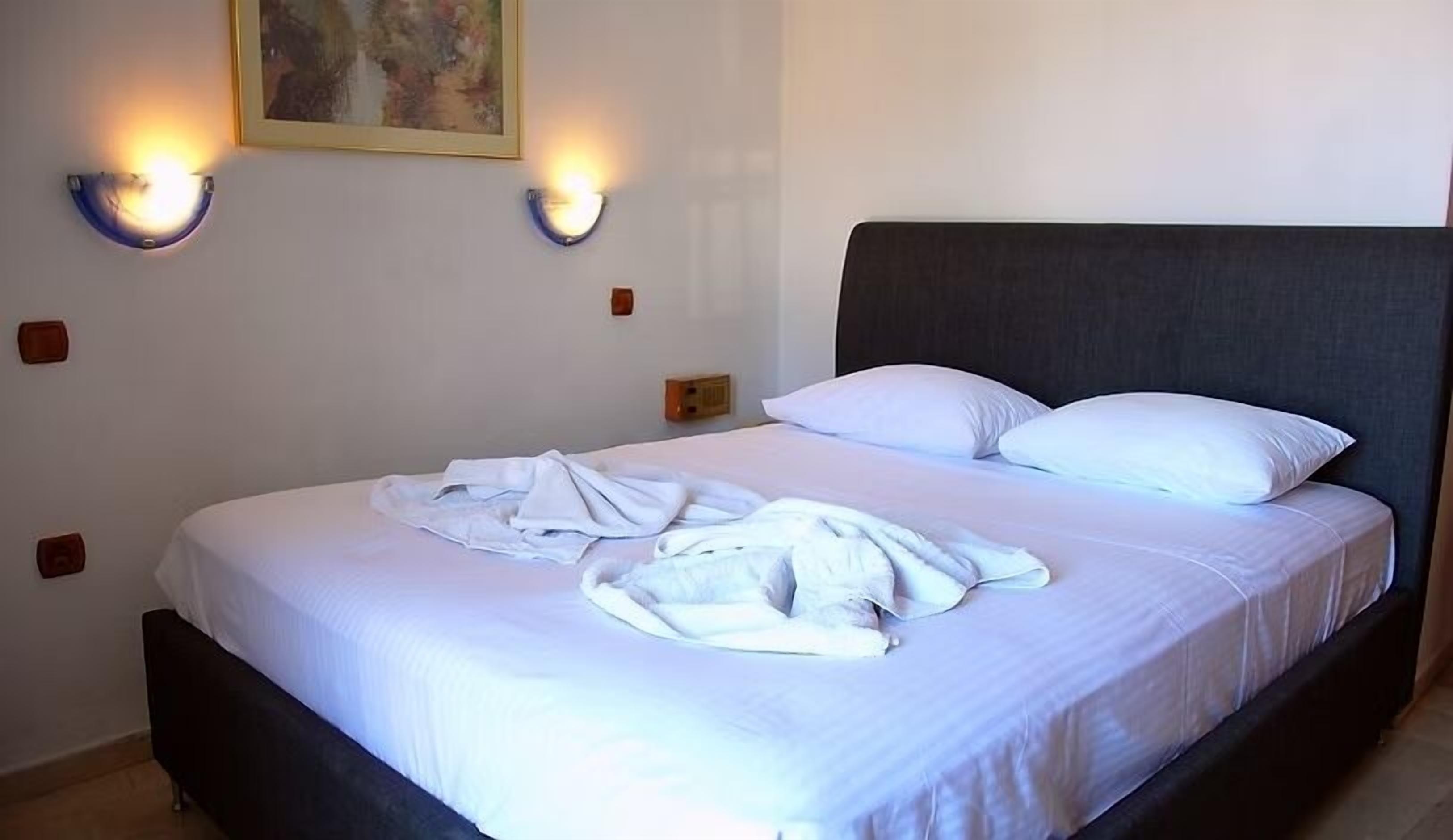 Una cama bien hecha con sábanas y almohadas blancas, dos lámparas de pared y un cuadro en la pared.