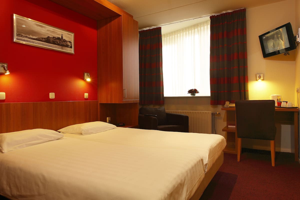 Habitación de hotel con cama, escritorio, silla, televisor y ventana con cortinas.