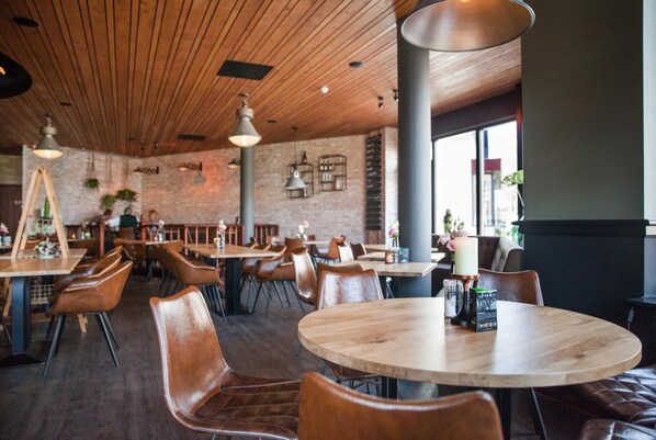A restaurant interior with wooden tables and chairs, brick walls, and hanging pendant lights.
