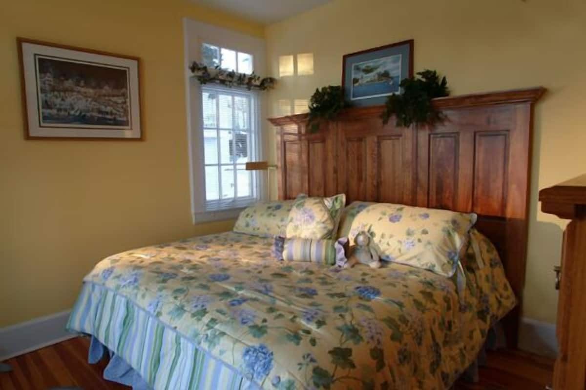 A bedroom with a floral bedspread, wooden headboard, and a window with a view.