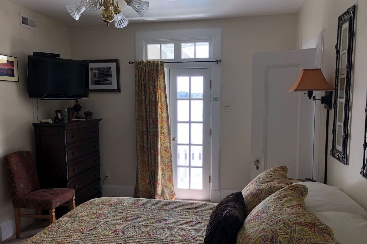 A bedroom with a bed, dresser, chair, television, and a window with curtains.