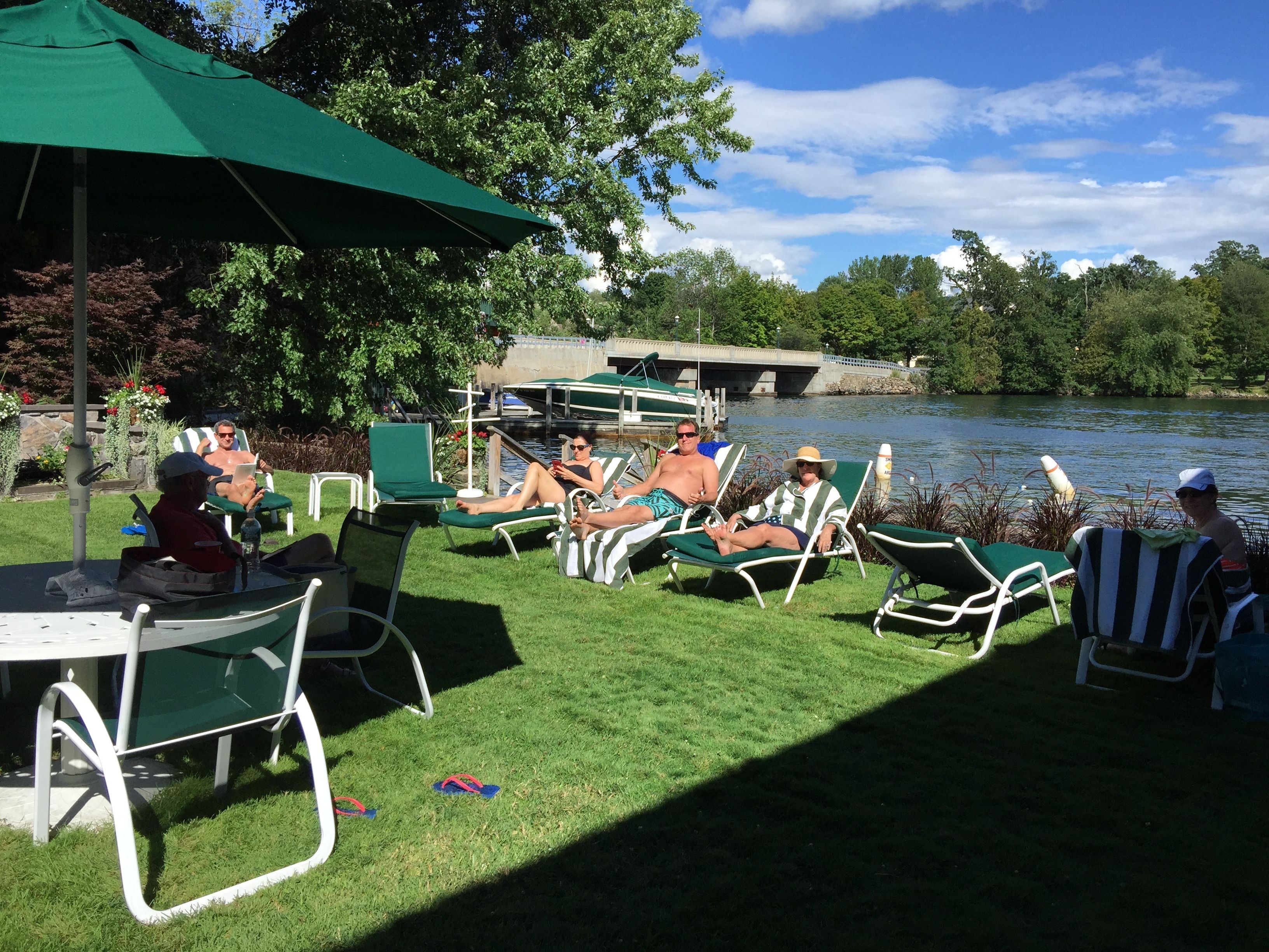 A lakeside patio with green lawn chairs and umbrellas, a dining table, and a view of the lake and trees.