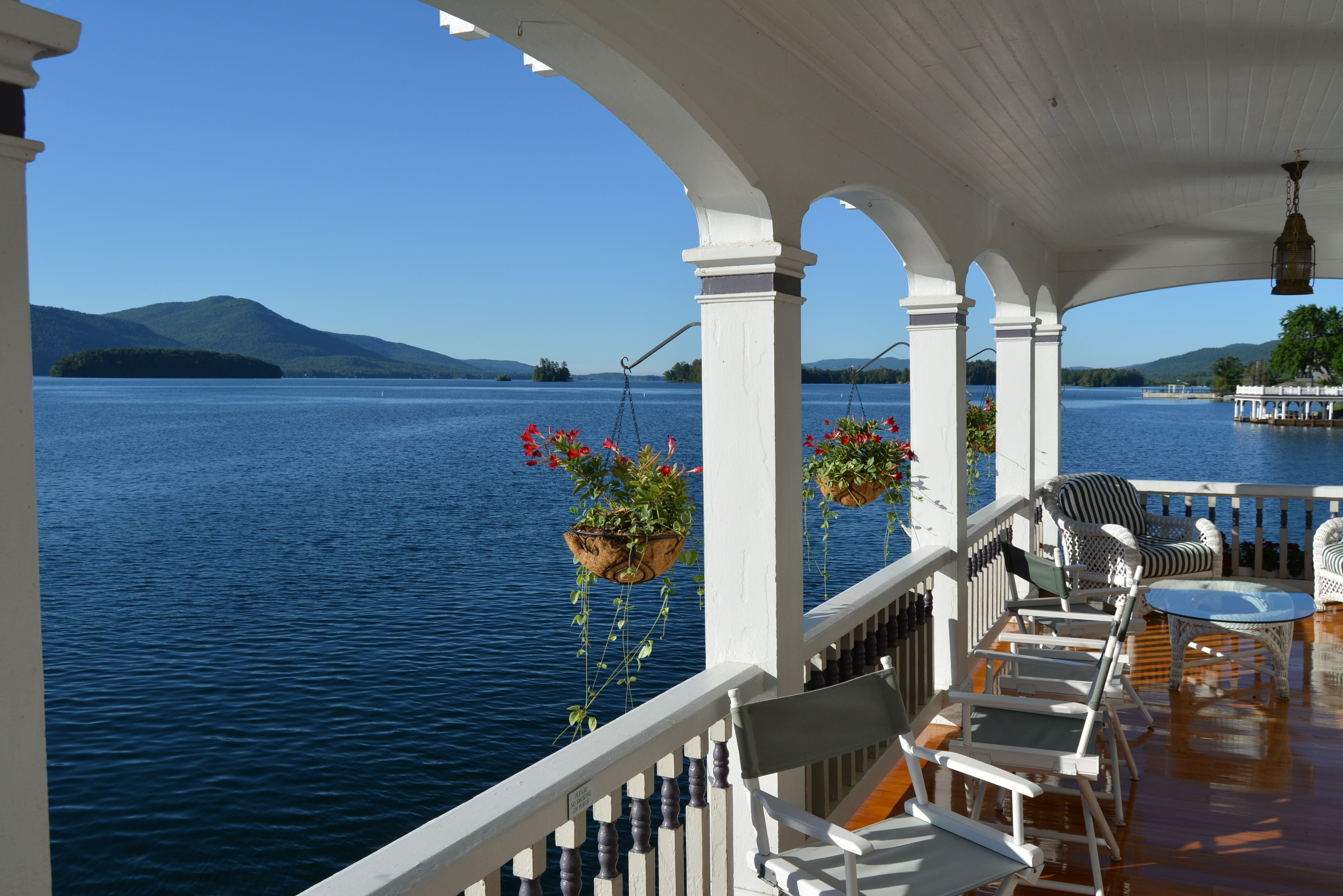 A covered outdoor area with a view of a lake and mountains. There are white columns, a railing, and outdoor furniture including chairs and a table.