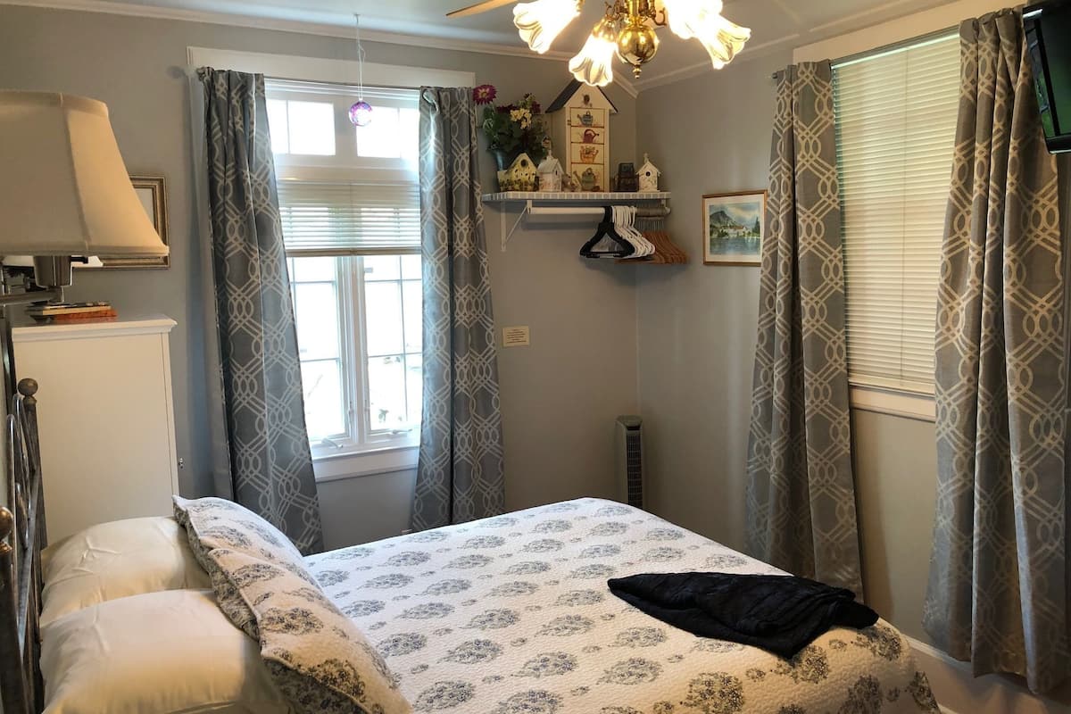A bedroom with a bed, a nightstand, a lamp, a window with curtains, a ceiling fan, and a wall-mounted shelf with decorative items.