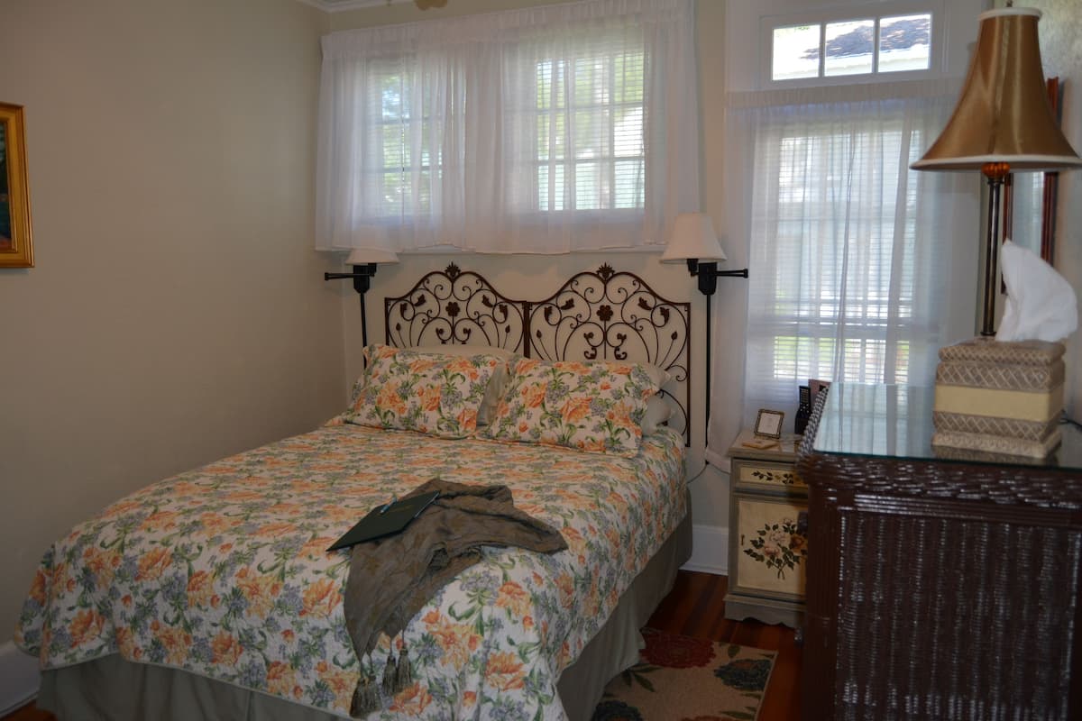 A bedroom with a floral bedspread, a wooden headboard, a nightstand with a lamp, and a chair.