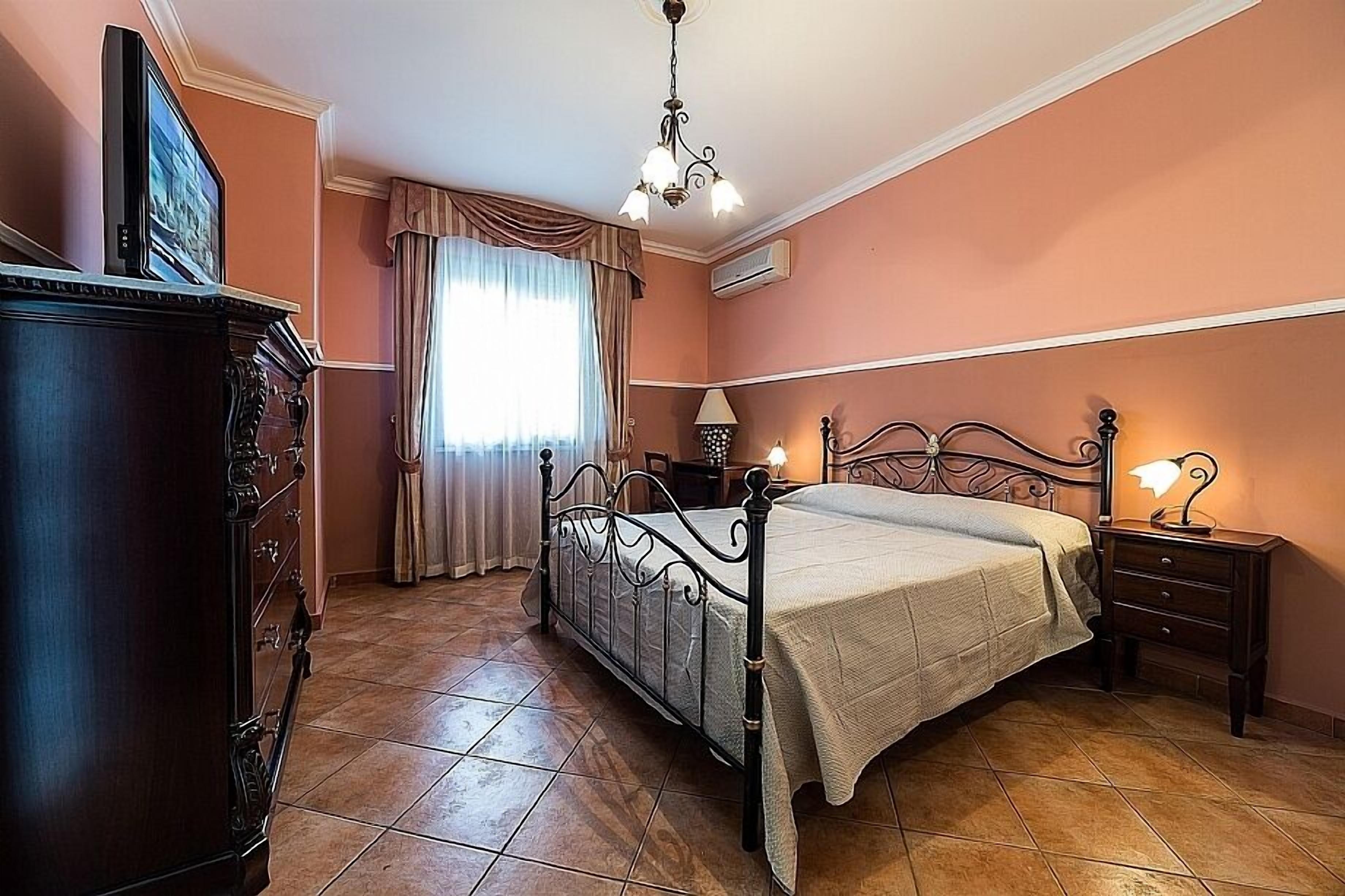 A bedroom with a bed, a dresser, a nightstand with a lamp, and a window with curtains.