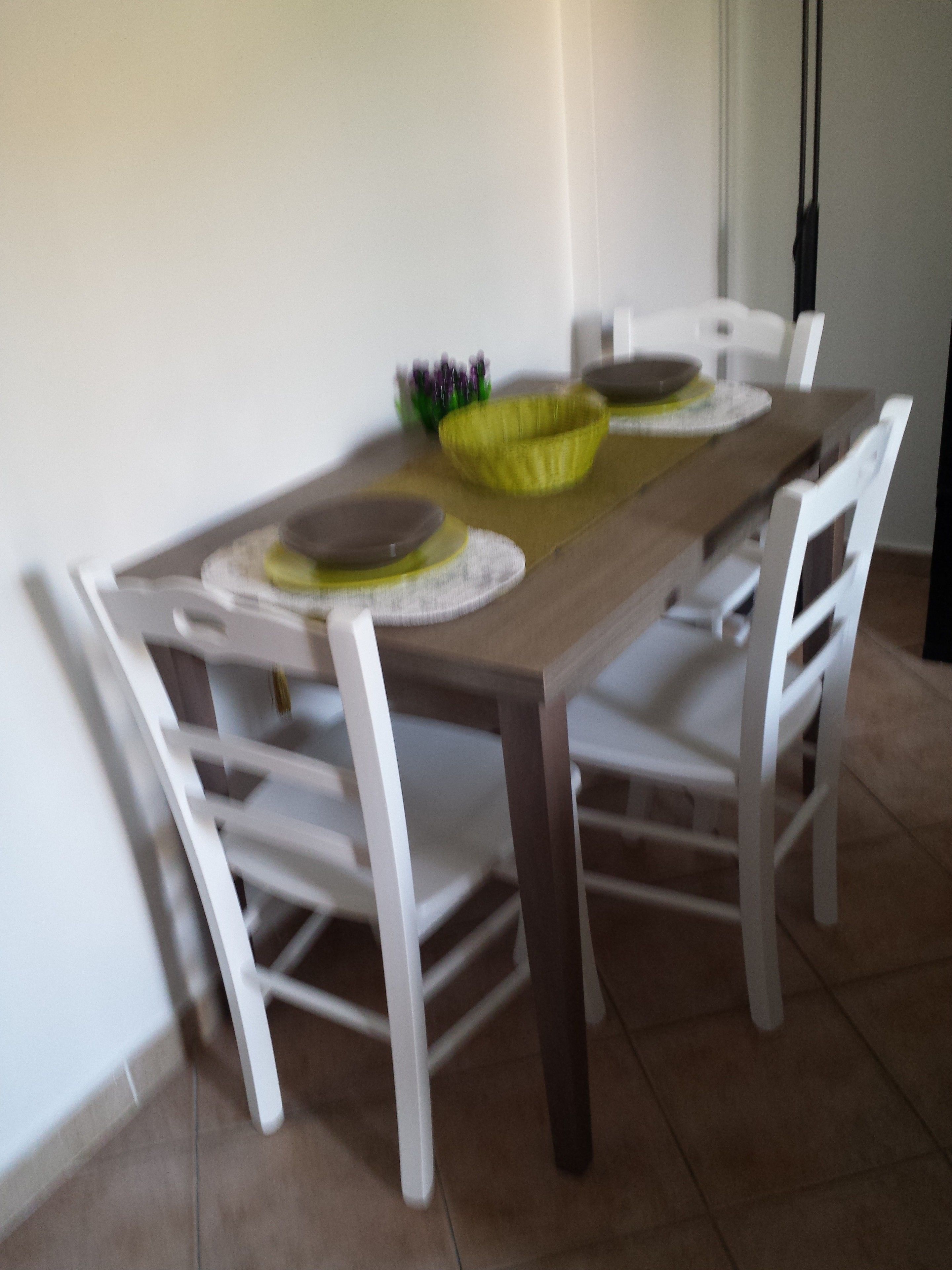Een eettafel met witte stoelen, een gele placemat en een schaal paarse bloemen.