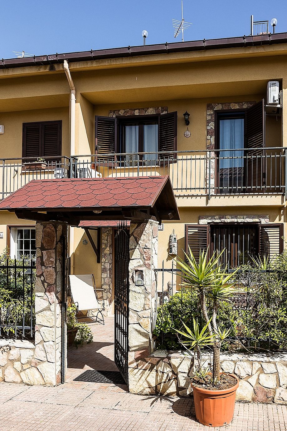 Un edificio con fachada de piedra, un balcón con barandilla metálica y una pequeña zona cubierta con una silla.