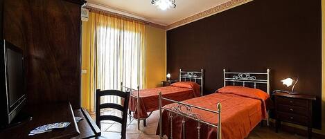 Habitación de hotel con dos camas, un escritorio con televisor, una silla, una mesita de noche con lámpara y una ventana con cortinas.