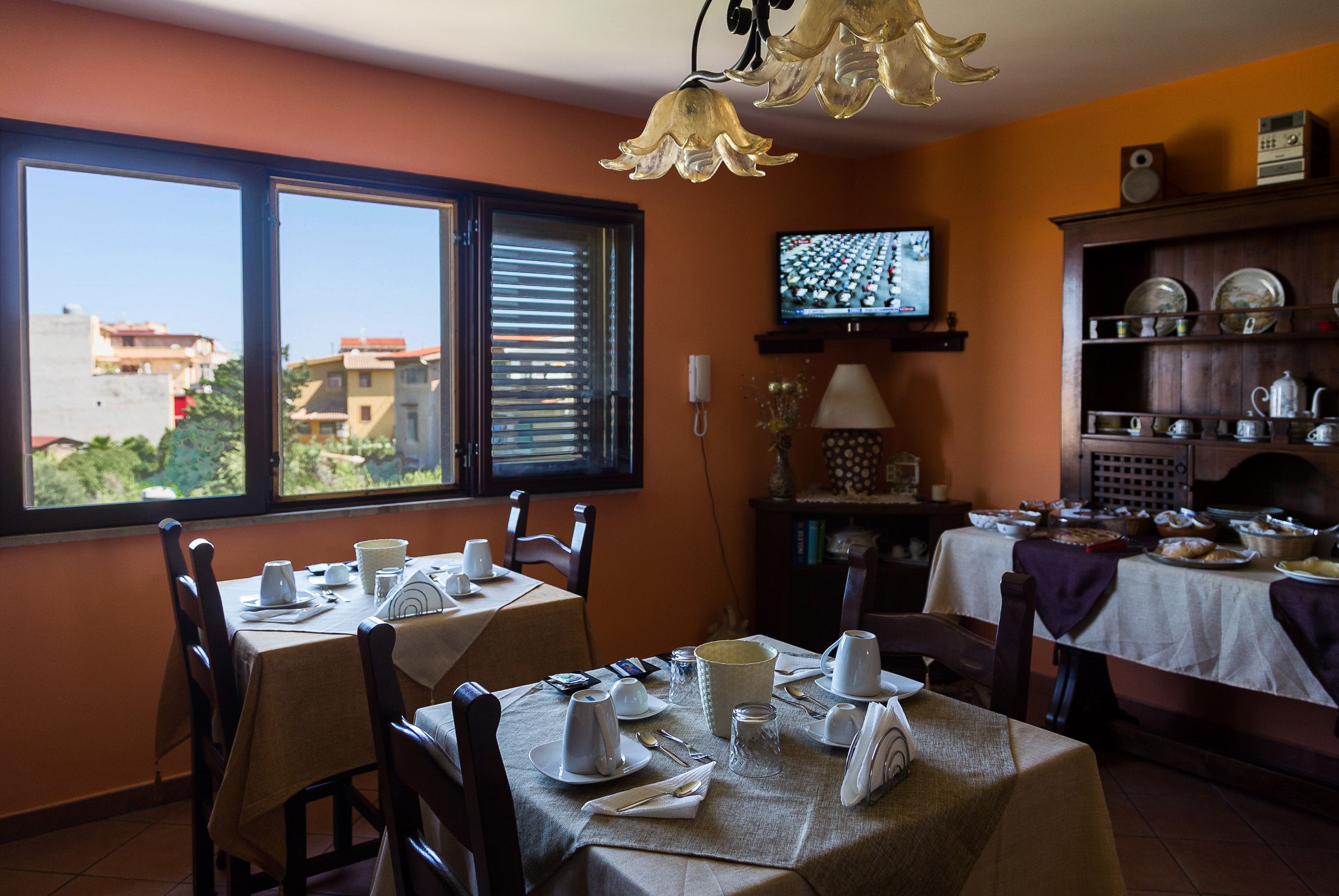Un comedor con mesas dispuestas para el desayuno, un televisor en la pared y vistas a edificios.