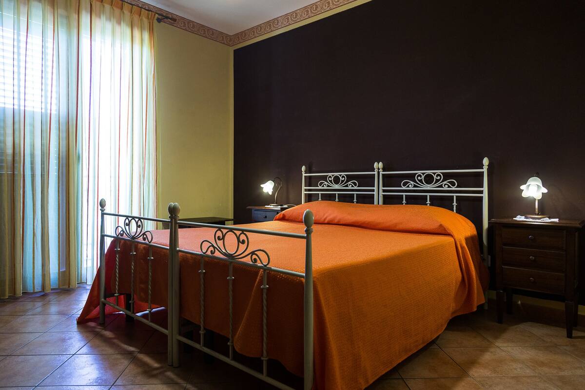 A bedroom with a metal bed frame, an orange bedspread, two bedside lamps, a nightstand, and a window with curtains.