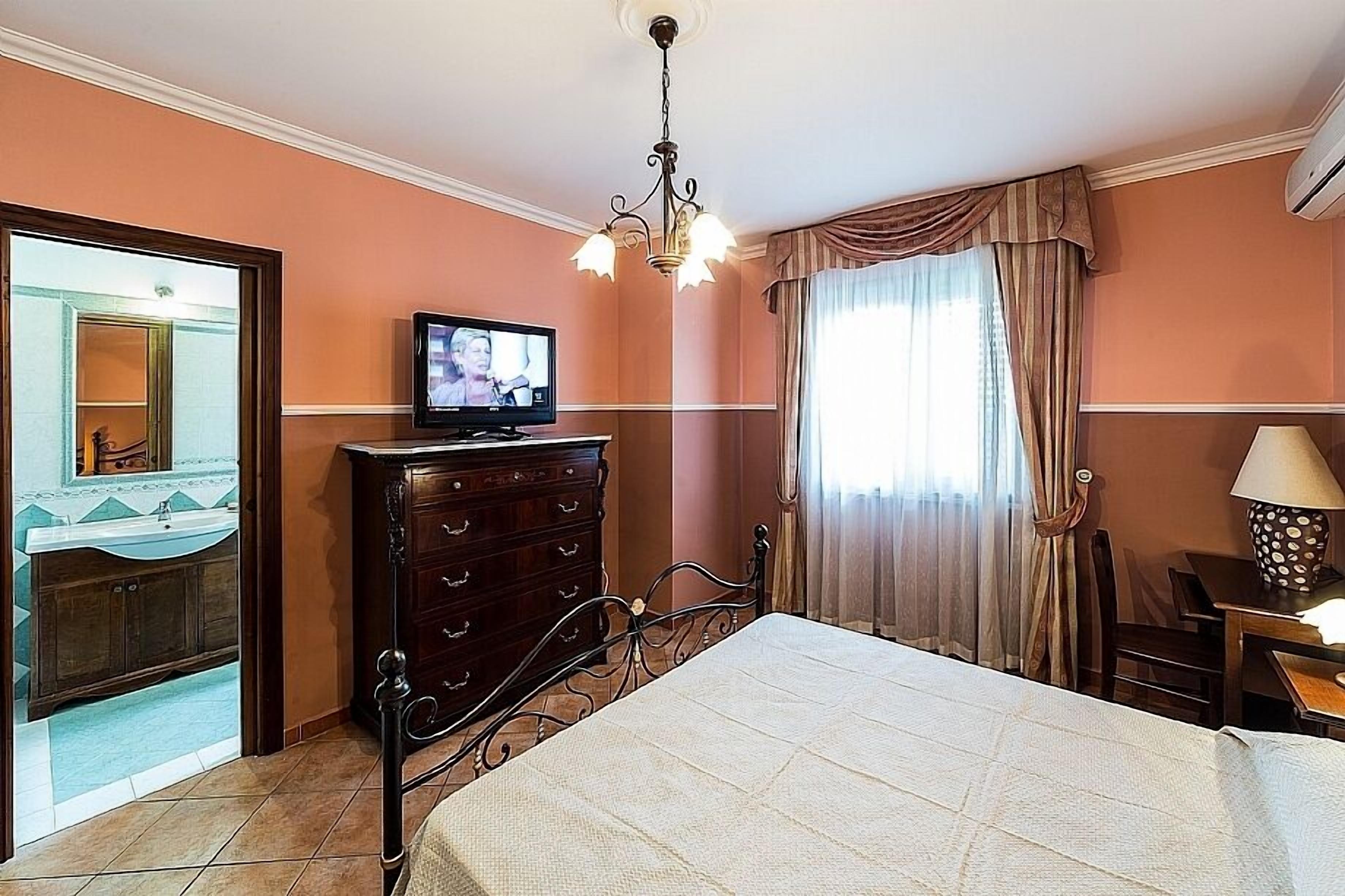 Ein Schlafzimmer mit einem Bett, einem Schminktisch, einem Fernseher, einer Kristallleuchter und einem Fenster mit Vorhängen.