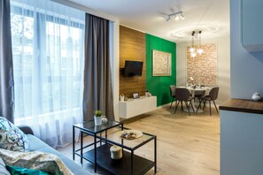 A modern living room with a sofa, a glass coffee table, a dining area with a table and chairs, a TV mounted on a wooden wall, and a green accent wall. - FriendHouse Apartments - Wawel Old City (Kraków)