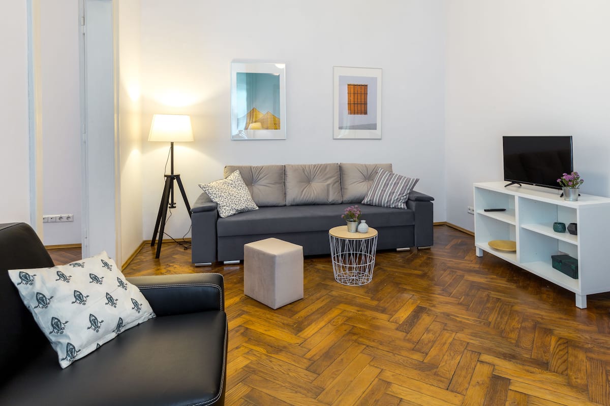 Ein modernes Wohnzimmer mit einem grauen Sofa, einem schwarzen Ledersessel, einem runden Beistelltisch mit Lampe und einem Flachbildfernseher auf einem weißen Lowboard.