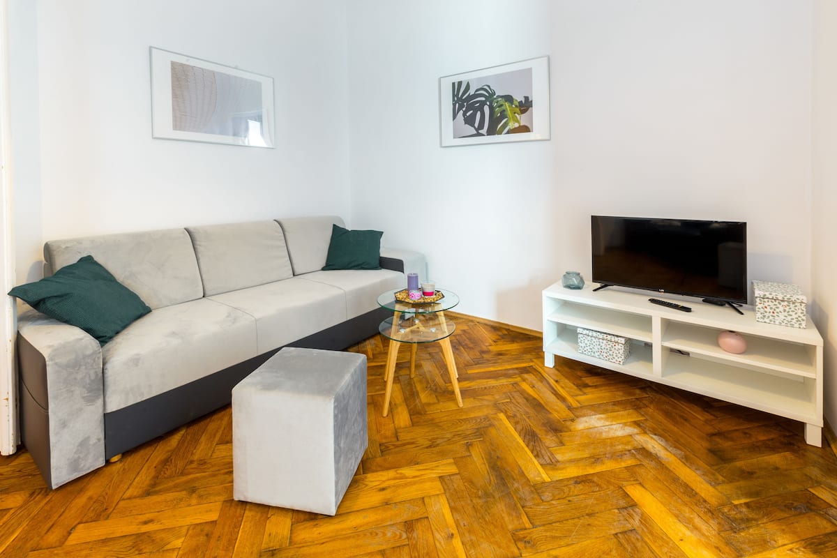 Ein modernes Wohnzimmer mit Sofa, Hocker, Glastisch, Fernseher und einem weißen Schrank.