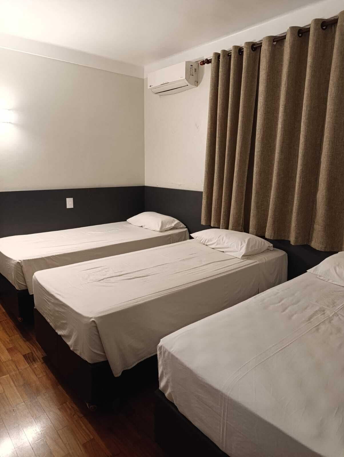 Habitación de hotel con tres camas individuales, piso de madera, aire acondicionado de pared y una cortina.