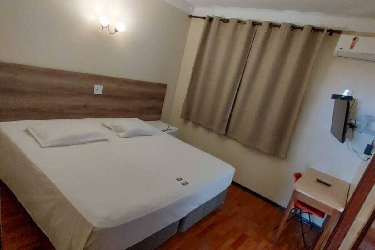 Habitación de hotel con cama, una mesita, una silla, ventana con cortinas y televisor montado en la pared.
