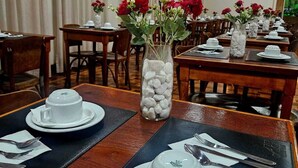Un espace repas avec des tables dressées pour deux personnes, chacune agrémentée d’assiettes blanches, de tasses et de couverts. Des vases avec des fleurs rouges et un grand vase rempli de galets blancs sont posés sur les tables.