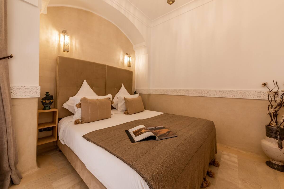 A neatly made bed with a brown blanket, a bedside table with a lamp, and a decorative vase on the floor.