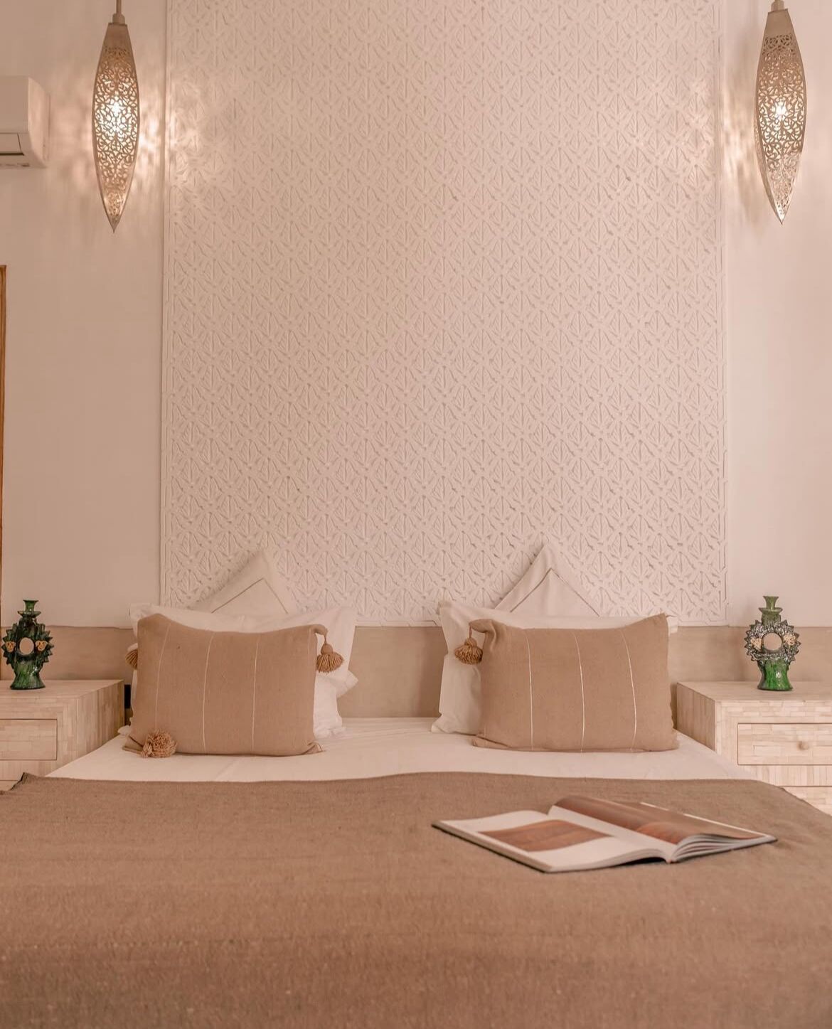 Une chambre avec un mur à motifs, deux lampes de chevet, un lit avec une literie beige et un livre posé sur le lit.