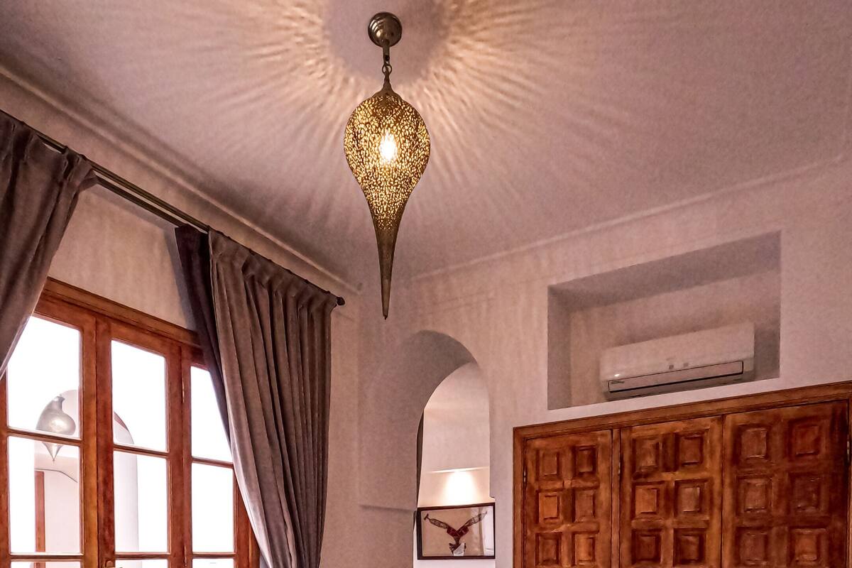 A bedroom with a large bed, a wooden wardrobe, a hanging light fixture, and a view through a window with curtains.