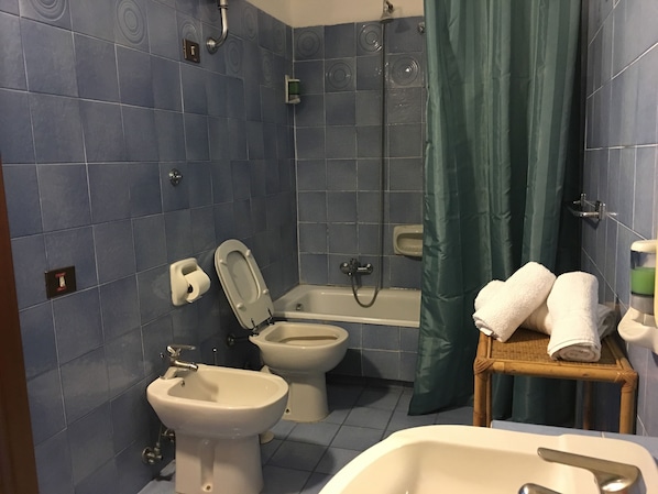Un baño con azulejos azules, una bañera, ducha, bidé e inodoro. Hay toallas sobre un taburete de madera.