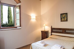 A bedroom with a bed, a bedside table with a lamp, a window with a view of trees, and a framed picture on the wall.