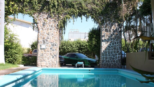 A swimming pool with stone pillars, a green car parked nearby, and a building in the background. - Hotel Yori (Cajeme)