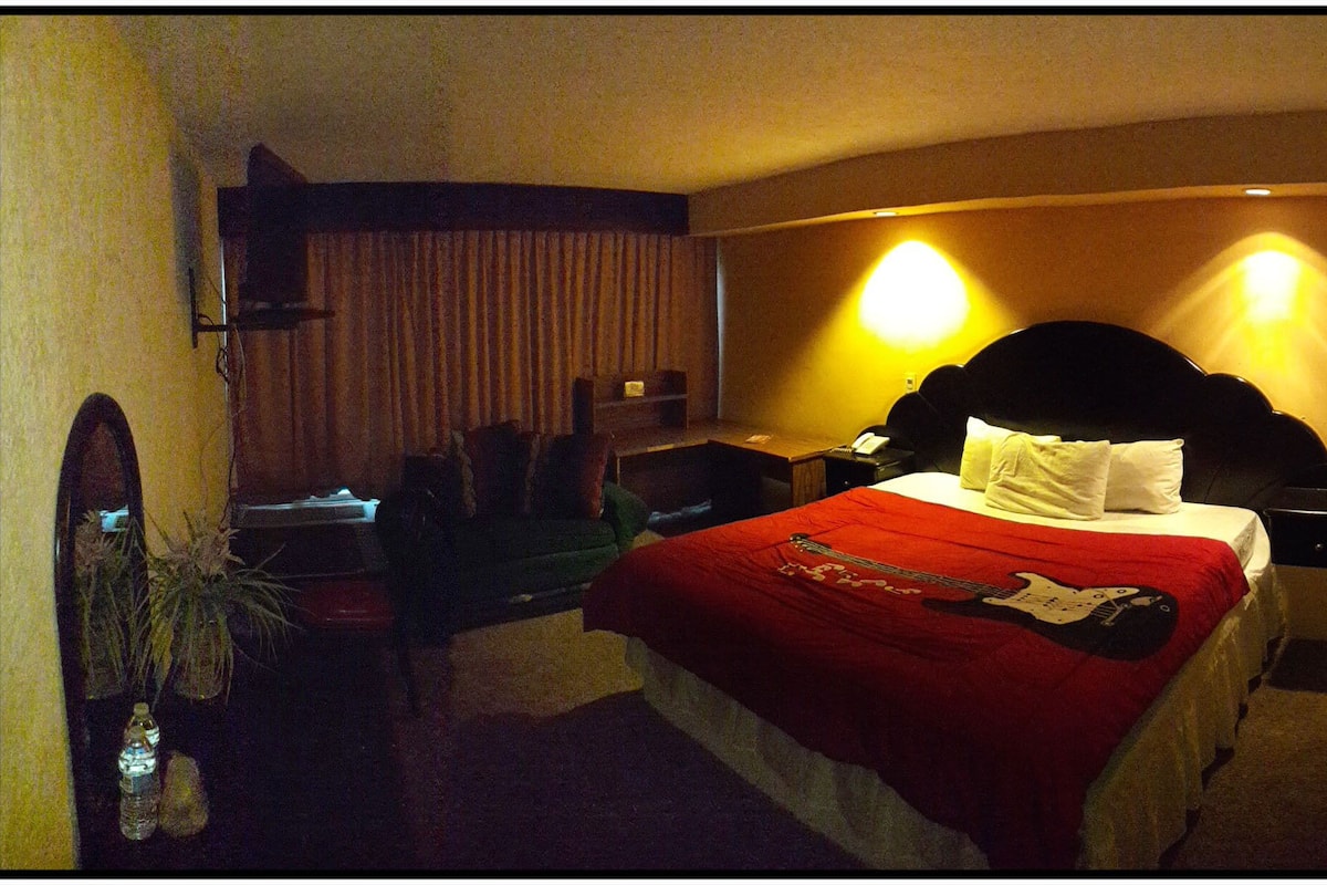 A hotel room with a bed, bedside table, lamp, and a small desk with a chair.