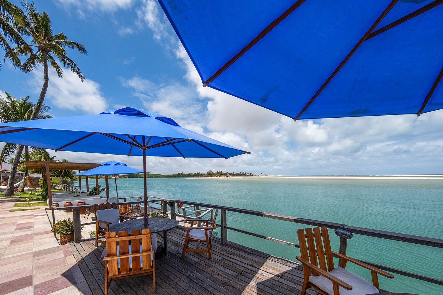 A wooden deck with outdoor seating, blue umbrellas, and a view of the ocean.