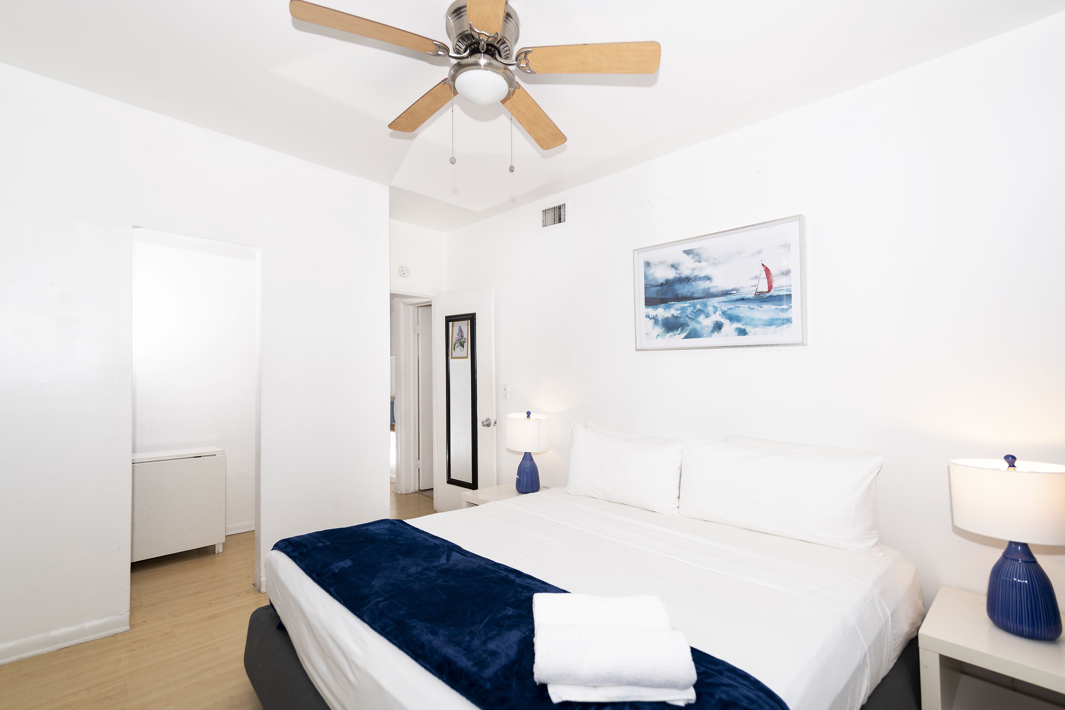 Een hotelkamer met een bed, een plafondventilator, een nachtkastje met een lamp, een schilderij aan de muur en een deur die naar een andere kamer leidt.