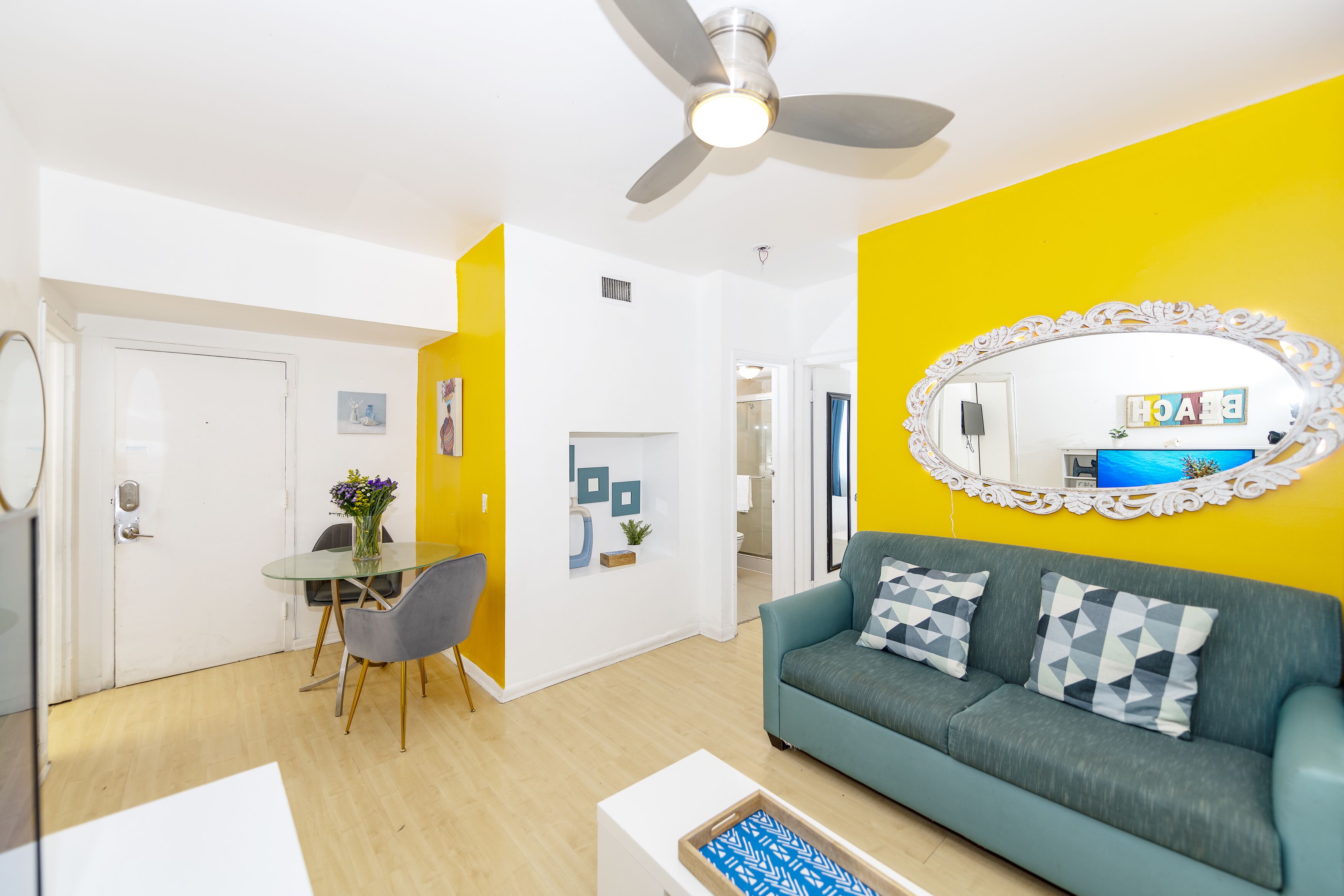 Ein Wohnzimmer mit einer Couch, einem Esstisch mit Stühlen, einem Deckenventilator und einem Spiegel an der Wand.
