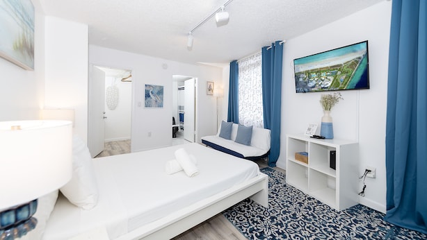 A modern hotel room with a bed, a desk, a chair, a TV, and a view of a coastal area. - Art Deco Ocean Walk Apartments (Miami Beach)
