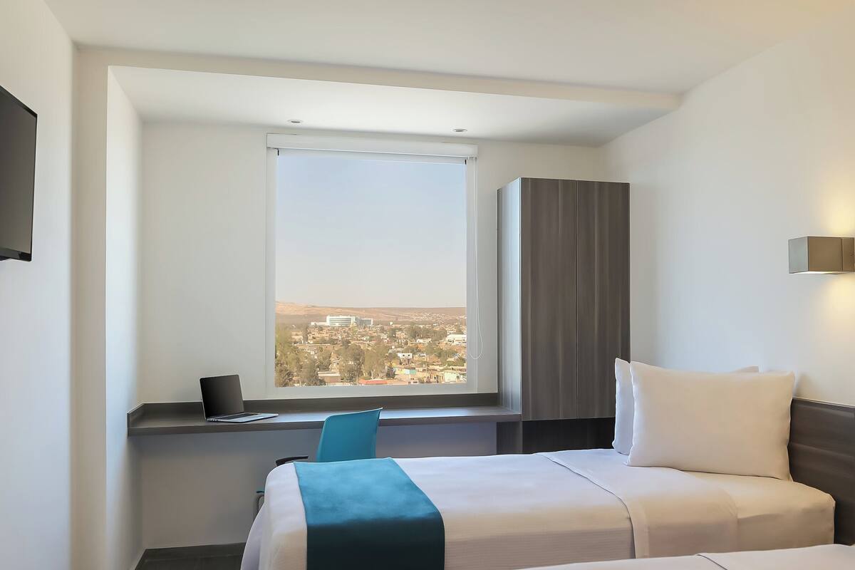 A hotel room with two beds, a desk with a laptop, a window with a city view, and a wardrobe.