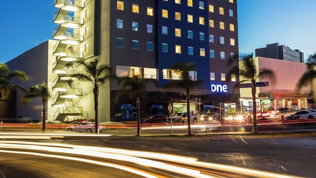 Un hôtel à plusieurs étages arborant la marque « One », caractérisé par un design moderne, des fenêtres illuminées et un panneau « One » bien visible sur la façade.