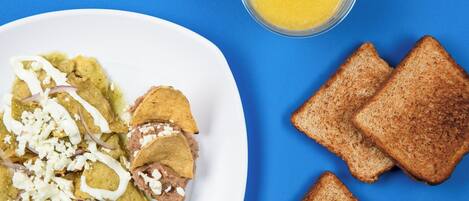 Un plato de nachos con queso y cebolla roja, acompañado de rebanadas de tostadas y un vaso de jugo de naranja sobre un fondo azul.