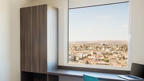 Une chambre à coucher avec un lit, un bureau avec un ordinateur portable, une chaise et une vue sur le paysage urbain par la fenêtre.
