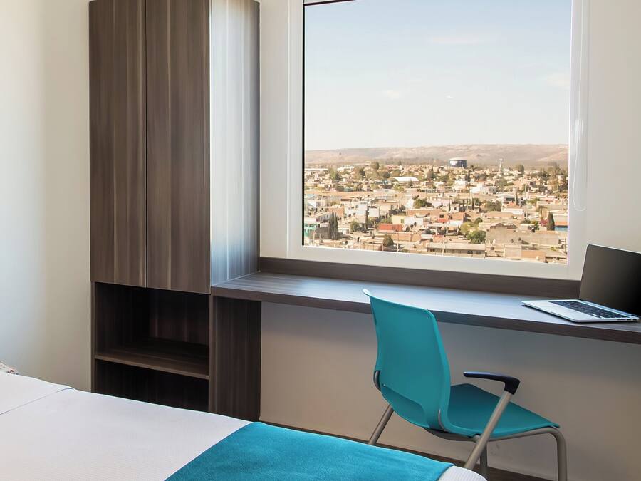 Une chambre à coucher avec un lit, un bureau avec un ordinateur portable, une chaise et une vue sur le paysage urbain par la fenêtre.