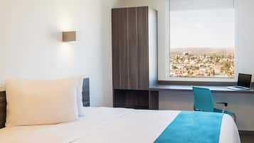 A hotel room with a bed, a desk with a laptop, a view of a cityscape, and a wardrobe.