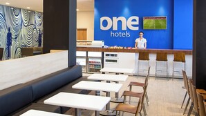 Un hall d’hôtel moderne avec un mur bleu orné du logo de la chaîne « One Hotels », un comptoir d’accueil, un espace salon et un comptoir de restauration.