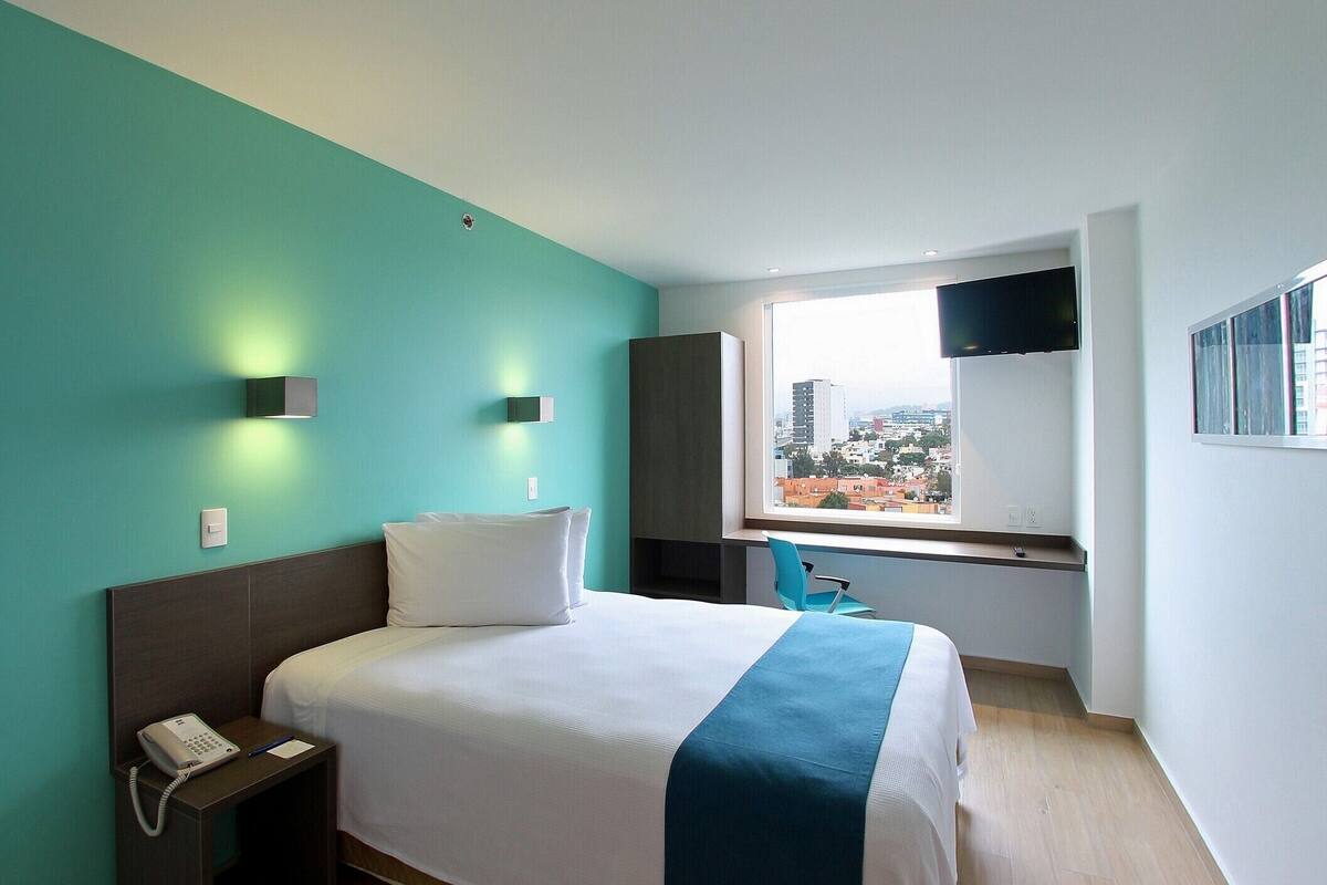 A hotel room with a bed, a desk, a chair, a phone, and a window with a city view.