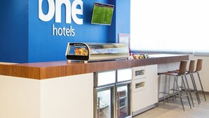 Un espace d’accueil avec un mur bleu orné de l’enseigne « One Hotels », un écran numérique diffusant un événement sportif, un buffet où les clients peuvent se servir eux-mêmes, et un coin salon avec des chaises.