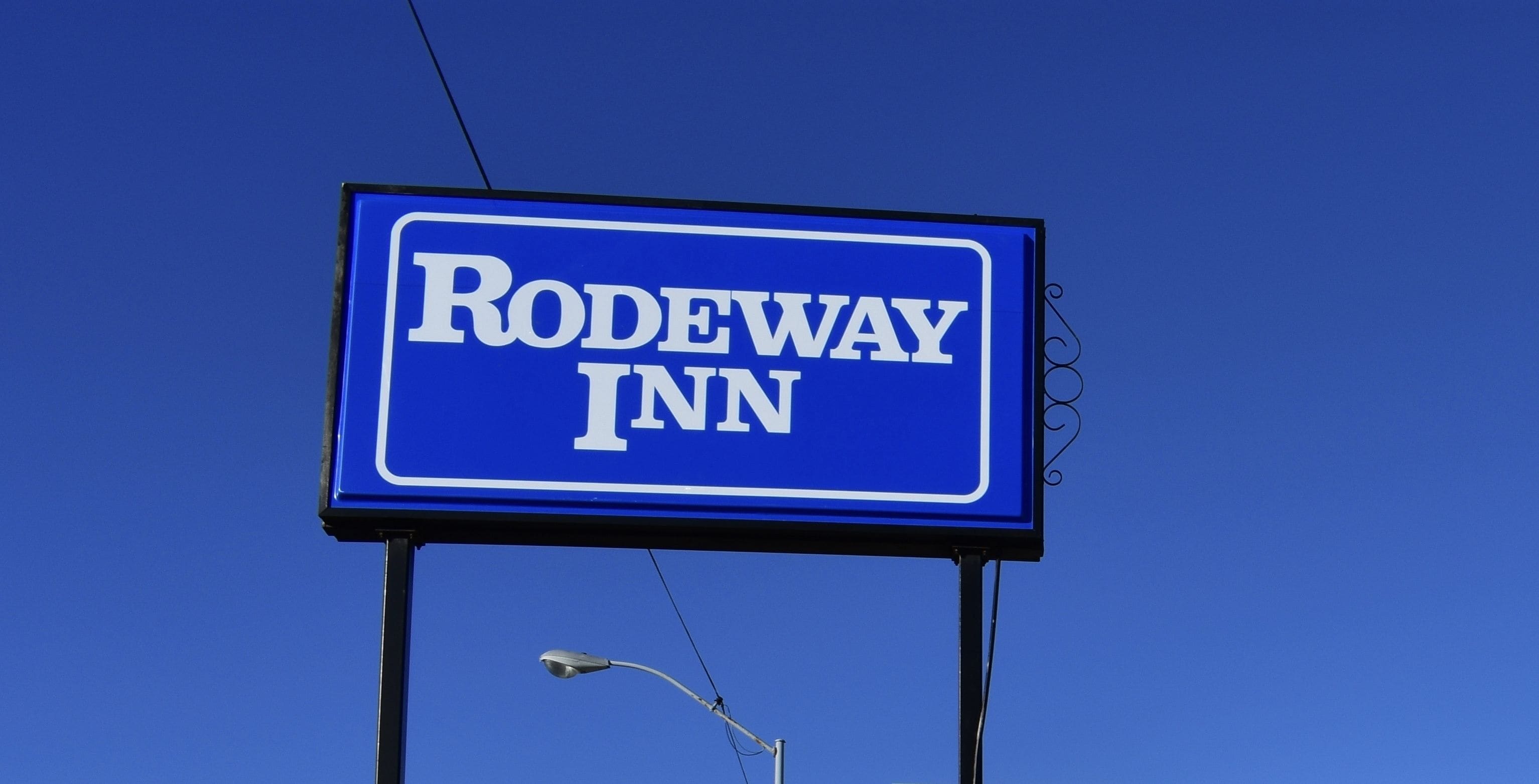 A blue sign with the text "RODEWAY INN" against a clear sky.